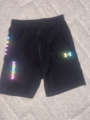 Svarta shorts från Under Armour - Säljer ett par svarta shorts från Under Armour med regnbågsfärgade loggor och text på ena benet. Shortsen har snörning i midjan och är perfekta för träning eller chill. Materialet känns lätt och andas bra.