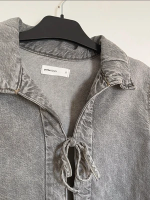 Grå jeansjacka med knytning - Snygg grå jeansjacka från Perfect Jeans med två knytband framtill istället för knappar. Jackan har klassisk krage, långa ärmar och en croppad passform. Perfekt för dig som vill ha en unik twist på den klassiska jeansjackan.