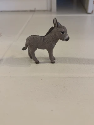 Grå åsna i plast - Säljer en liten åsna i plast, perfekt som dekoration eller leksak. Den är grå med svart man och svans, samt vita detaljer runt nosen och ögonen. Figuren har realistiska former och står stadigt på fyra ben.