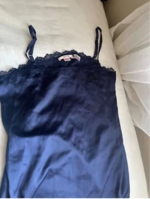 Mörkblått nattlinne med spetsdetaljer - Snyggt mörkblått nattlinne i satin med tunna axelband och spetskant upptill. Perfekt för en bekväm och stilren look på kvällen. Materialet är glansigt och mjukt mot huden. Axelbanden är justerbara för bästa passform.