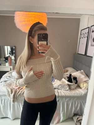 Beige off shoulder stickad tröja - Trendig beige stickad tröja med off shoulder-modell och kabelstickat mönster. Långa ärmar med breda ribbade muddar och snygg passform som framhäver midjan. Perfekt för en stilren och mysig look.