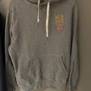 Grå hoodie från Morris med tryck - Grå hoodie från Morris med guldfärgat emblem och brittisk flagga broderat på bröstet. Tröjan har vit snörning i huvan och en stor magficka framtill. Mjuk bomullskänsla och klassisk passform, perfekt för chill dagar.
