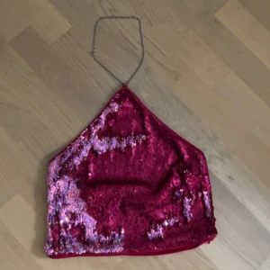 Glittrig rosa paljett halterneck topp - Säljer en glittrig röd halterneck topp med paljetter och kedja runt halsen. Toppen har en trekantig form och är croppad, perfekt för att sticka ut på festen. Framsidan är täckt av röda och rosa paljetter, medan baksidan är slät röd tyg. Supersnygg att styla med jeans eller kjol!