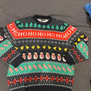 Jultröja H&M XL med snögubbar och granar - Säljer en färgglad jultröja från H&M i storlek XL. Tröjan har mönster med snögubbar, julgranar, klockor och texten HO HO HO. Färgerna är svart, röd, grön, gul och vit. Tillverkad i 100% återvunnen polyester och har rund hals och lång ärm.