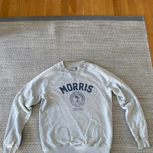 Grå sweatshirt från Morris - Snygg grå sweatshirt från Morris med blå logga och tryck framtill. Klassisk rund halsringning och ribbade muddar vid ärmslut och nederkant. Perfekt för en chill och stilren look.