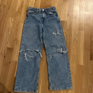 Blå ripped jeans från H&M - Säljer ett par blå jeans från H&M med höga midja och raka ben. Byxorna har flera stora slitningar och hål på framsidan för en edgy look. Klassisk femficksmodell med knapp och dragkedja.