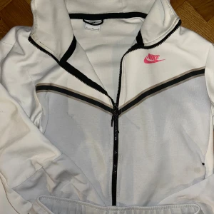 Nike Tech Vit Rosa  - Nike hoodie i vit med svarta och grå detaljer, hel dragkedja och huva. Rosa Nike-logga på bröstet ger en snygg kontrast. Tröjan har långa ärmar och sportig design, perfekt för chill eller träning. Materialet är mjukt och känns skönt mot huden.
