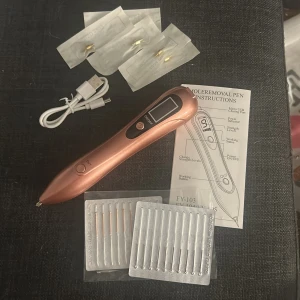 Mole Removal Pen FY-103 - Elektrisk mole removal pen i kopparfärg med LCD-display, flera styrkenivåer och tillbehör. Inkluderar laddkabel, nålar och instruktioner. Produkten ser ut att vara i mycket gott skick utan synligt slitage. Perfekt för hudvård hemma. Aldrig använd