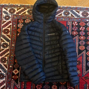 Svart dunjacka Peak Performance - Säljer en svart dunjacka från Peak Performance med huva och dragkedja. Jackan är quiltad och har en stilren design med logga på bröstet. Perfekt för kalla dagar och har en snygg, normal passform. Lagad på ärmen, syns på bild 3! Annars i bra skick 