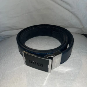 Svart skärp från Hugo Boss i skinn - Snyggt svart skärp från Hugo Boss i skinn med stilren, rektangulär metallspänne där det står BOSS. Bältet har en klassisk design med diskreta blå sömmar och passar perfekt till jeans eller kostymbyxor. Metallspännet har en modern känsla och ger ett exklusivt intryck.