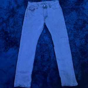 Levi's 501 ljusblå jeans straight fit - Ljusgrå Levi's 501 jeans med klassisk straight fit.  Jeansen har knappgylf, brunt läderpatch bak och den ikoniska röda Levi's-taggen på bakfickan. Tillverkade i slitstarkt bomullsjeansmaterial, perfekta för en chill och tidlös look. Obs ett litet hål börjar komma fram under gylfen se bild 4