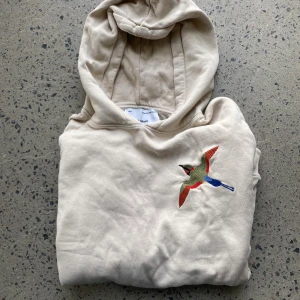 Beige hoodie från Axel Arigato med broderad fågel - Snygg beige hoodie från Axel Arigato med broderad färgglad fågel på bröstet. Tröjan har huva, känguruficka och ribbade muddar. Perfekt för dig som gillar stilrena plagg med en cool detalj.