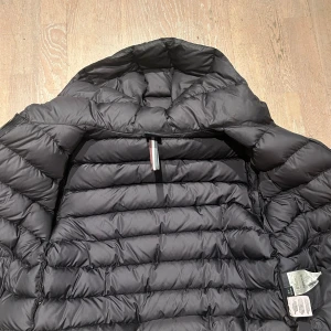 Svart dunjacka från Colmar - Säljer en svart dunjacka från Colmar med klassisk puffer-design och huva. Jackan har horisontella sömmar, dragkedja och en snygg logotyp på ärmen. Perfekt för kalla dagar när du vill hålla stilen och värmen.