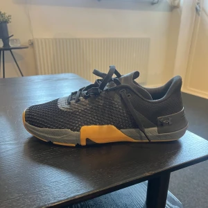 Svarta Under Armour träningssneakers - Säljer ett par svarta Under Armour sneakers med mesh-ovandel och snygga detaljer i grått och orange. Skorna har snörning och en platt, slitstark sula som är perfekt för gymmet. Snygg sportig look och bekväm passform för dig som gillar att träna. Säljes på grund av fel storlek, aldrig använda.