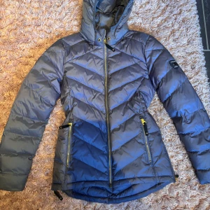 Grå dunjacka från Canada Snow - Snygg grå/blå dunjacka från Canada Snow med huva och gulddetaljer på dragkedjorna. Jackan har ett quiltat mönster i sicksack, två fickor med dragkedja och logga på ärmen. Jackan kostar 2200kr i nypris. Använd 1/2 gånger