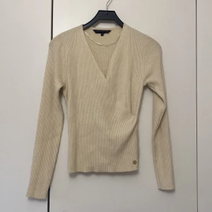 Beige omlottopp Pernilla Wahlgren S - Trendig beige omlottopp från Pernilla Wahlgren Design. Ribbstickad med smal passform, långa ärmar och en liten gulddetalj nertill. Perfekt för dig som gillar stilrena och enkla plagg med en twist.