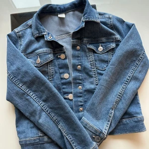 Klassisk blå jeansjacka från 8Denim - Snygg blå jeansjacka från 8Denim med klassisk passform, silverfärgade knappar och två bröstfickor med lock. Jackan är tillverkad i slitstarkt denim och har långa ärmar samt stilrena sömmar. Perfekt för dig som vill ha en tidlös look.