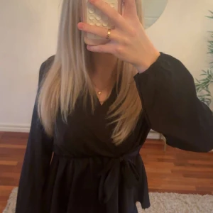 Svart blus - Svart blus med långa ärmar och knytband i midjan. Blusen har en v-ringad halsringning och ett flowy fall som ger en snygg siluett. Perfekt för dig som gillar stilrena och enkla plagg med en twist.