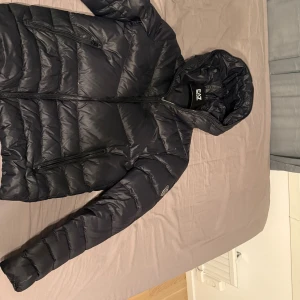 Svart pufferjacka från EA7 Emporio Armani - Snygg svart pufferjacka från EA7 Emporio Armani med huva och dragkedja framtill. Jackan har två sidofickor med dragkedja och en stilren logga på ärmen. Perfekt för kalla dagar och ger en clean look. Jackan har ett väldigt litet hål men går såklart att fixa för biligt.