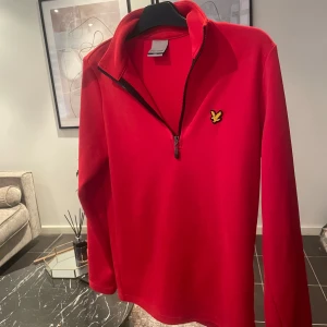Röd half zip från Lyle & Scott - Riktigt snygg röd långärmad tröja från Lyle & Scott med gul logga på bröstet. Tröjan har half zip-dragkedja och hög krage, perfekt för lager-på-lager. Mjuk och skön polyester som passar till både chill och streetwear.