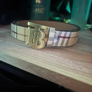 Burberry bälte med guldspänne - Säljer ett snyggt Burberry bälte med klassiskt rutigt mönster i beige, rött och svart. Bältet har ett stort guldigt metallspänne med Burberrys ikoniska TB-logga. Perfekt accessoar för att lyfta din outfit.