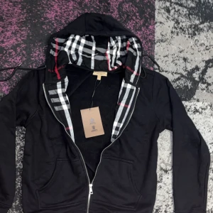 Svart hoodie från Burberry med rutig huva - Svart hoodie från Burberry med klassisk rutig insida i huvan och längs dragkedjan. Tröjan har huva med snörning och dragkedja framtill. Perfekt för dig som vill ha en stilren men ändå ikonisk look.    Står att storleken är L men M i verkligheten🚨