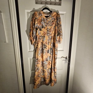 Blommig långklänning - Superfin långklänning med aprikos/ljusrosa botten och blommigt mönster. Klänningen har trekvartsärmar med resår, knappar framtill och en avslappnad passform. Perfekt för dig som gillar en romantisk och trendig stil.