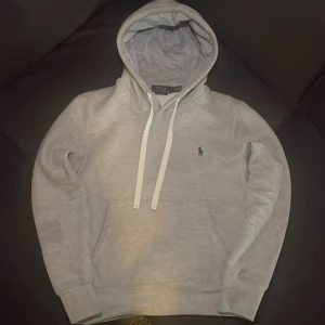 Grå hoodie från Polo Ralph Lauren - Snygg grå hoodie från Polo Ralph Lauren i storlek S. Tröjan har huva med vita snören, lång ärm och klassisk mörkblå broderad logga på bröstet. Mjuk bomullskvalitet och ribbade muddar vid ärmslut och nederkant. Perfekt för chill dagar och streetstyle.