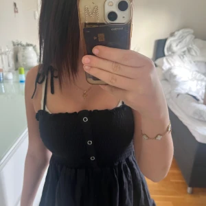 Svart topp med knytband - Supersöt svart topp med smockad överdel, knappar framtill och knytband på axlarna. perfekt för varma dagar. Enkel och stilren design som är lätt att matcha med accessoarer.