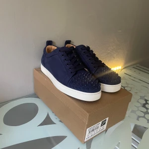 Mörkblå Christian Louboutin sneakers - Säljer ett par mörkblå Christian Louboutin sneakers i mocka med coola nitar på tån och klassisk röd sula. Skorna har snörning och vit platt sula, och kommer med originalkartong och dustbag. Perfekt för dig som vill sticka ut med exklusiva detaljer.