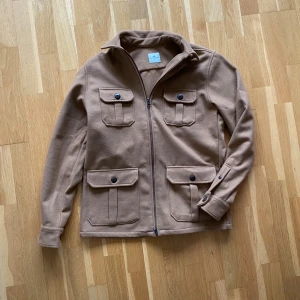 Beige overshirt från John Henric (helt ny) - Snygg beige overshirt från John Henric med fyra stora fickor framtill och stilrena knappar. Jackan har dragkedja och klassisk krage, perfekt att slänga på över en hoodie. Materialet är mjukt och känns lyxigt, passar dig som gillar clean och enkel stil. (Aldrig använd) nypris på ca 1,100 ‼️pris kan såklart diskuteras ‼️