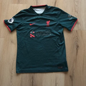 Liverpool FC Elliott 19 - Snygg mörkgrön Liverpool FC fotbollströja från Nike med Elliott och nummer 19 på ryggen. Kortärmad med röda detaljer vid ärmslut och krage, Premier League patch på ärmen och Dri-FIT material som andas. Diskret mönster på tyget och klubbmärke på bröstet.