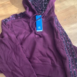 Vinröd hoodie med leopardmönster Adidas - Säljer en vinröd hoodie från Adidas med leopardmönstrad insida på huvan och längs axlarna. Tröjan har snörning vid huvan, klassiska Adidas-ränder och broderad logga på bröstet. Materialet är mjuk bomull och polyester, perfekt för dig som gillar streetstyle.