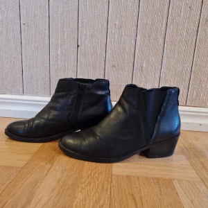 Svarta stövlar i äkta läder strl 38 - Snygga svarta stövlar i äkta läder med låg klack och rund tå. Skorna har dragkedja på insidan och elastiska partier på sidorna för enkel påtagning. Storlek 38.