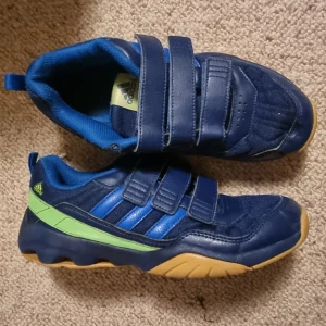 Blå sneakers från Adidas med kardborre - Säljer ett par blå innesneakers från Adidas med gröna detaljer och gula sulor. Skorna har tre kardborreband för enkel påtagning och en sportig look. Ovandelen är i syntet och textil, perfekt för dig som gillar färg och bekvämlighet.
