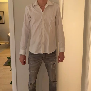 Vit skjorta från Bläck, tailored fit - Klassisk vit skjorta från Bläck i storlek L med snygg tailored fit. Skjortan har lång ärm, krage och knappar framtill. Perfekt för dig som gillar stilrena och enkla plagg. Materialet är mjukt och känns som bomull, vilket gör den skön att bära hela dagen.