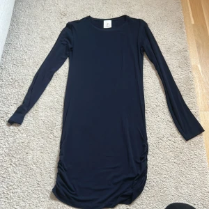 Mörkblå långärmad klänning - En stilren mörkblå klänning med lång ärm och rund halsringning. Modellen är rak och har lätt rynkade detaljer i sidorna nedtill för en snygg siluett. 