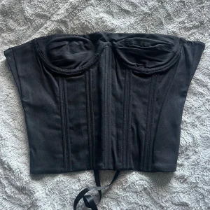 Svart korsett med snörning - Säljer en svart korsett med snygga kupor och markerade sömmar framtill. Baksidan har snörning med svarta satinband som ger en justerbar passform. Materialet känns stadigt och har en matt finish. Perfekt för att skapa en edgy look.