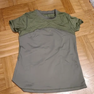 Grön sportig t-shirt i mesh XS - Snygg grön t-shirt i sportig stil med meshdetaljer upptill och slätare tyg nedtill. Perfekt för träning eller aktiva dagar. Tillverkad i 73% polyester och 27% spandex för stretch och komfort. Kortärmad och med rund hals.