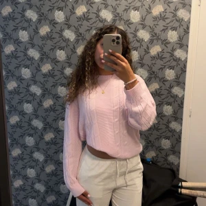 Ljusrosa croppad stickad tröja - Säljer en ljusrosa croppad stickad tröja med kabelstickat mönster och långa ärmar. Tröjan har en relaxed fit och ribbade muddar vid ärmslut och nederkant. Perfekt för dig som gillar en söt och trendig stil.