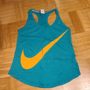 Turkos Nike linnetopp med orange logo - Säljer en turkos linnetopp från Nike i storlek XS. Toppen har en stor orange Nike Swoosh framtill och klassisk brottarrygg. Perfekt för träning eller när du vill ha en sportig look. Tillverkad i mjuk bomull för skön känsla.