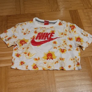 Nike croppad blommig kort t-shirt XS - Nike croppad t-shirt i vitt med stora blommiga mönster i gult och orange. T-shirten har korta ärmar och en stor röd Nike-logga framtill. Perfekt för dig som gillar färg och sportig stil.