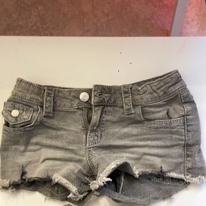 Grå jeansshorts från Gina Tricot - Snygga grå jeansshorts från Gina Tricot med fransig kant och slitna detaljer. Modellen har fem fickor, bälteshällor och stängs med knapp och dragkedja. Perfekt för dig som gillar en cool och avslappnad stil.