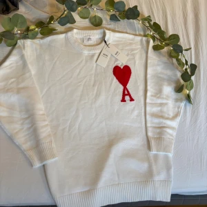 Vit stickad tröja från Ami Paris - Vit stickad tröja från Ami Paris med röd hjärtlogga och bokstaven A på bröstet. Tröjan har rund hals, ribbade muddar och är långärmad. Perfekt för dig som gillar stilrena plagg med en twist.