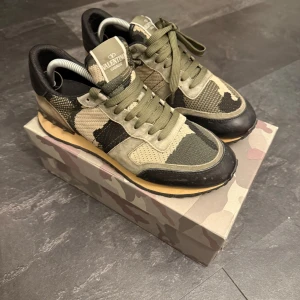 Valentino Rockrunner sneakers camouflage - Snygga Valentino Rockrunner sneakers i grönt, beige och svart med camouflage-mönster. Skorna har snörning, detaljer i mocka och skinn samt coola nitar på hälen. Perfekta för dig som vill sticka ut med exklusiva och trendiga sneakers. Kvitto kan lösas
