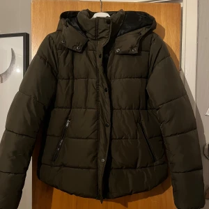 Mörkgrön pufferjacka med huva - Mörkgrön pufferjacka med huva och dragkedja framtill. Jackan har två snedställda fickor med dragkedja och tryckknappar längs öppningen. Perfekt för kalla dagar och riktigt skön att bära. Passformen är normal och jackan är vadderad för extra värme.