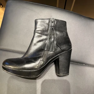 Svarta stövletter i skinn med klack vagabond - Säljer ett par svarta stövletter i skinn med chunky klack och rund tå. Skorna har dragkedja på sidan och snygga tofsdetaljer som ger en cool touch. Perfekta för dig som gillar en stilren men edgy look.