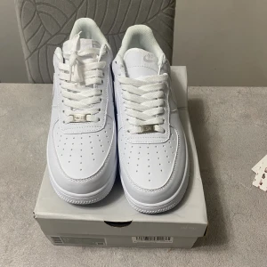 Nike Air Force 1 helvita sneakers - Klassiska Nike Air Force 1 sneakers i helvitt skinn med vit sula och snörning. Ikonisk design med perforerad tå och Nike-logga på sidan. Perfekta för dig som vill ha en clean och tidlös look. Lågt skaft och platt sula för maximal komfort.