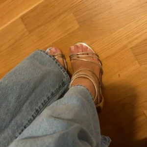 Guldiga sandaler med hög klack - Säljer ett par glansiga guldiga sandaler med smala remmar och öppen tå. Skorna har hög klack och dragkedja bak för enkel påtagning. Perfekta för dig som vill sticka ut med metallic vibes och elegant look.