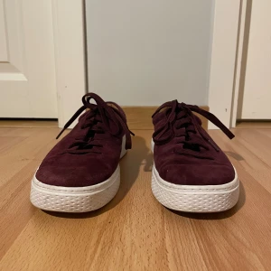 Vinröda Polo sneakers i mocka - Snygga vinröda sneakers från Polo med ovandel i mjuk mocka vit och röd platt sula. Skorna har klassisk snörning och rund tå. Polo-logga i guld baktill. 